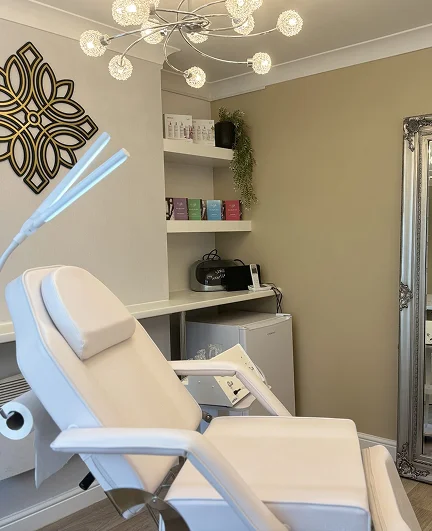 Modern beauty or treatment room with a white reclining chair, decorative lighting, shelves with skincare products, and a large ornate mirror.