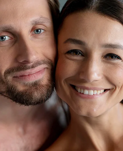 Close-up of a smiling bearded man with blue eyes leaning affectionately against a partner whose face is partially visible beside him.