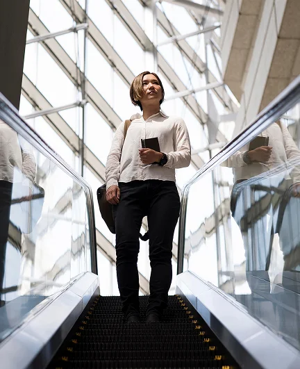 Person in business casual clothing riding down an escalator in a modern glass building, holding a phone and shoulder bag.
