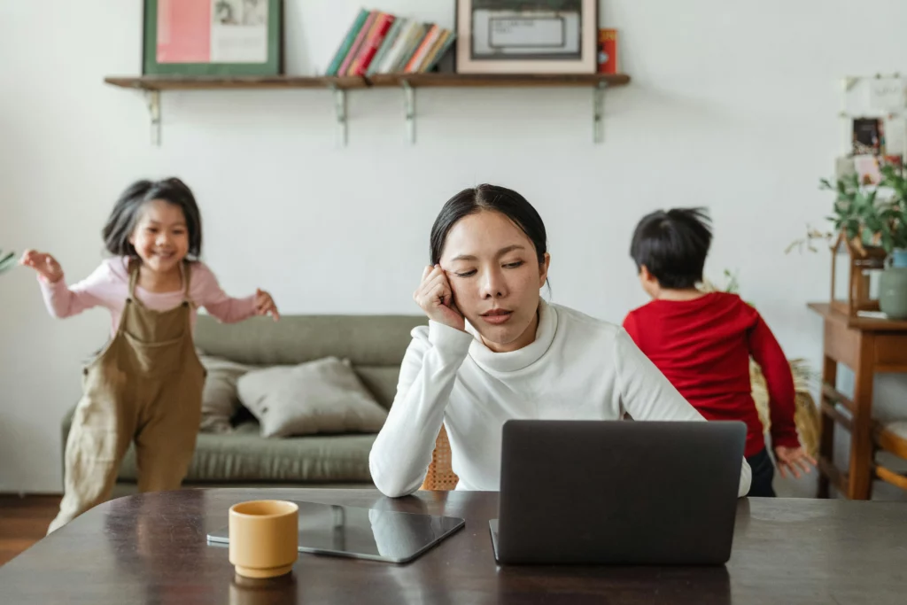 Stressed parent working on a laptop at a dining table while two young children play energetically in the background in a living room.