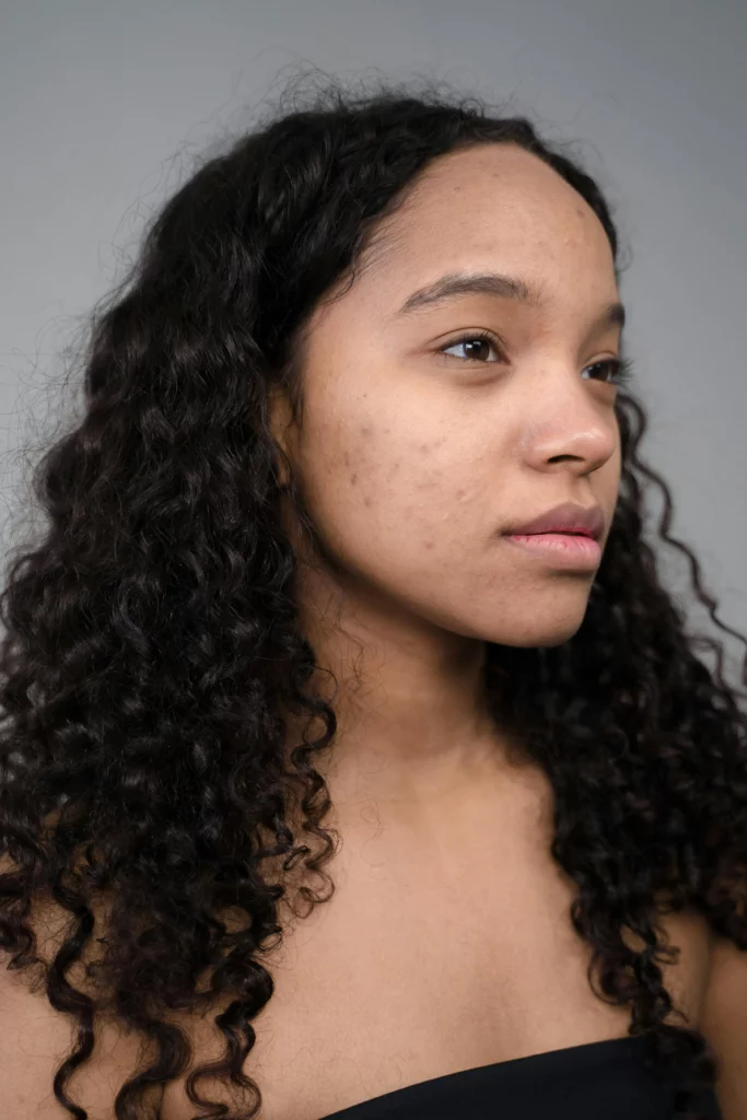 Close-up of a woman with long, dark, curly hair wearing a strapless black top against a plain neutral background.