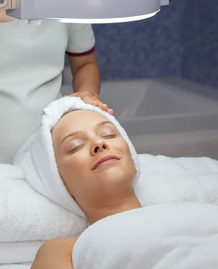 Esthetician performing a facial treatment on a client lying on a spa bed, with the client’s head wrapped in a white towel under a magnifying lamp.