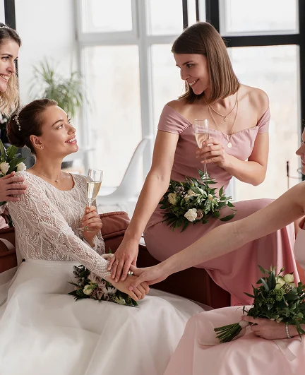 Bride and bridesmaids in pink dresses sitting together indoors, holding bouquets and champagne glasses while stacking their hands in a celebratory gesture.