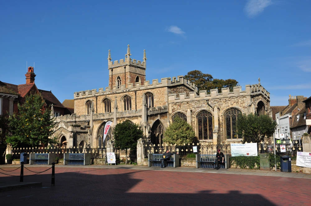 Historic stone church with crenelated roofline, arched stained-glass windows, and a central tower overlooking a paved town square on a sunny day.
