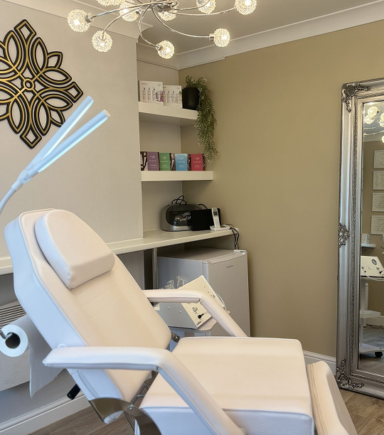 Modern beauty or treatment room with a white reclining chair, decorative lighting, shelves with skincare products, and a large ornate mirror.