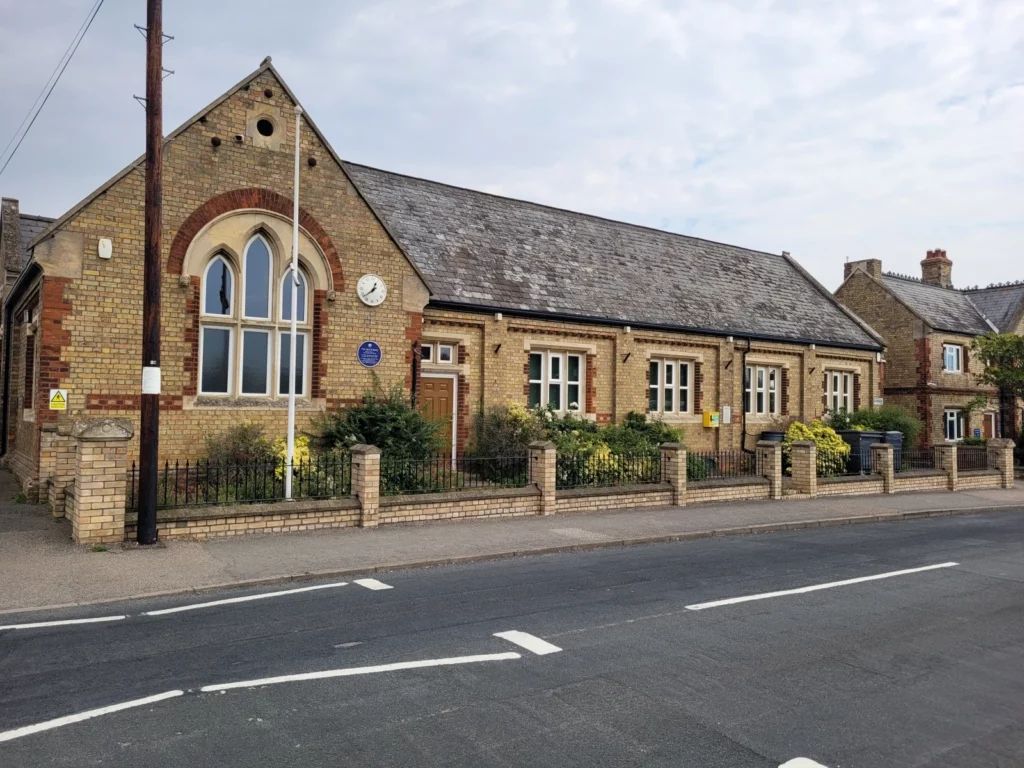Street view of a long, single-story brick building with arched windows, a slate roof, and a small clock on the facade, bordered by a low brick fence along the road.