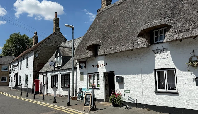 Street of traditional whitewashed cottages with thatched and tiled roofs, including a small business named “The Nook,” on a sunny day.