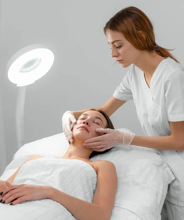 Esthetician in white scrubs gently examining or massaging a client’s face while the client lies on a treatment bed under a bright circular lamp.