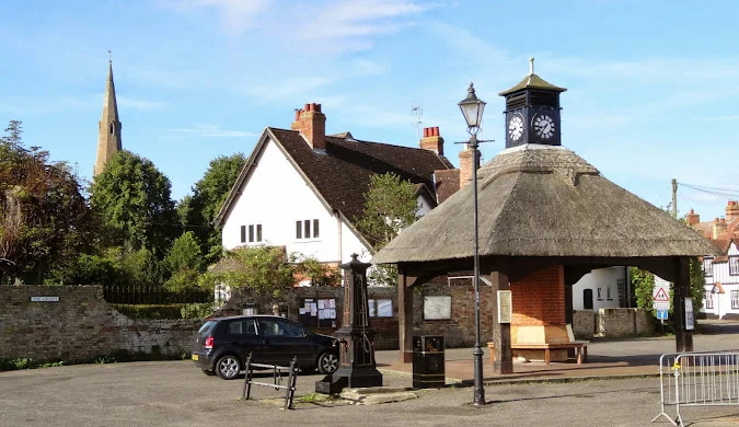 Quiet village square with a thatched-roof clock pavilion, benches, and a parked car, surrounded by traditional houses and a church spire in the background under a clear blue sky.