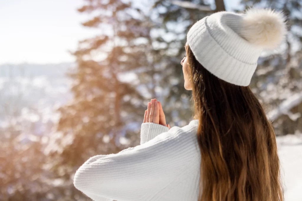 Woman in a white knit hat and sweater standing outdoors in a snowy forest, holding her hands together in a prayer or meditation pose while facing the winter sunlight.